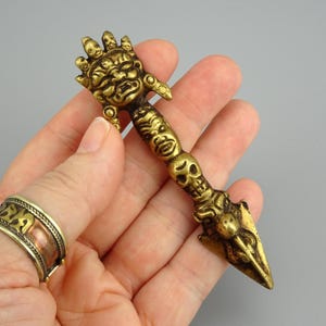 May include: A detailed brass ritual dagger with a hand-shaped top, faces, and a skull. The dagger is held in a hand, and a brass ring is visible on the finger. The dagger has a pointed end.