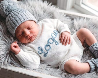 May include: A newborn baby wearing a light gray knit hat with a pom-pom, a white knit romper with the name "Logan" in blue, and matching gray knit booties. The baby is lying on a gray faux fur blanket inside a white wooden box.