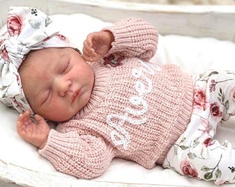 Może przedstawiać: Śpiące dziecko w różowym swetrze z napisem "Love" w białym skrypcie, kwiatowe spodnie i pasująca opaska na głowę w kwiaty. Dziecko leży w białej drewnianej kołysce.
