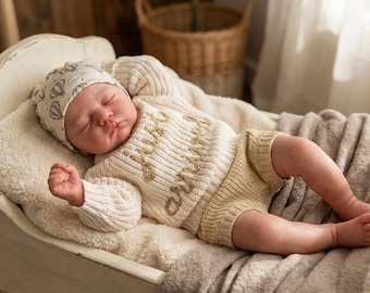 Könnte beinhalten: Ein schlafendes Baby in einem cremefarbenen Strickpullover und Shorts-Set mit den Worten "hello and" auf dem Pullover gestickt. Das Baby trägt auch einen passenden Hut mit einem Heißluftballonmuster. Das Baby liegt in einer weißen Holzwiege.