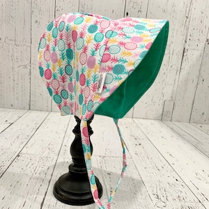 May include: A white sun hat with a pineapple print in pink, yellow, and turquoise. The hat has a green lining and ties at the back.