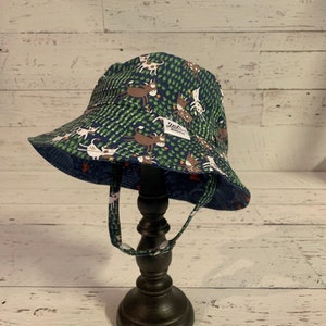 Op de afbeelding: Een groene en blauwe zonnehoed met een witte achtergrond met een patroon van cartoondieren. De hoed heeft een brede rand en een kinband.