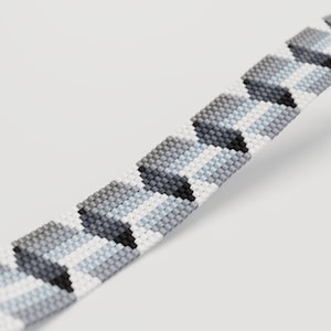 May include: A beaded bracelet featuring a geometric pattern in shades of gray, white, and black. The bracelet is made of small, evenly spaced beads, creating a textured surface. The design is a repeating pattern of shapes.