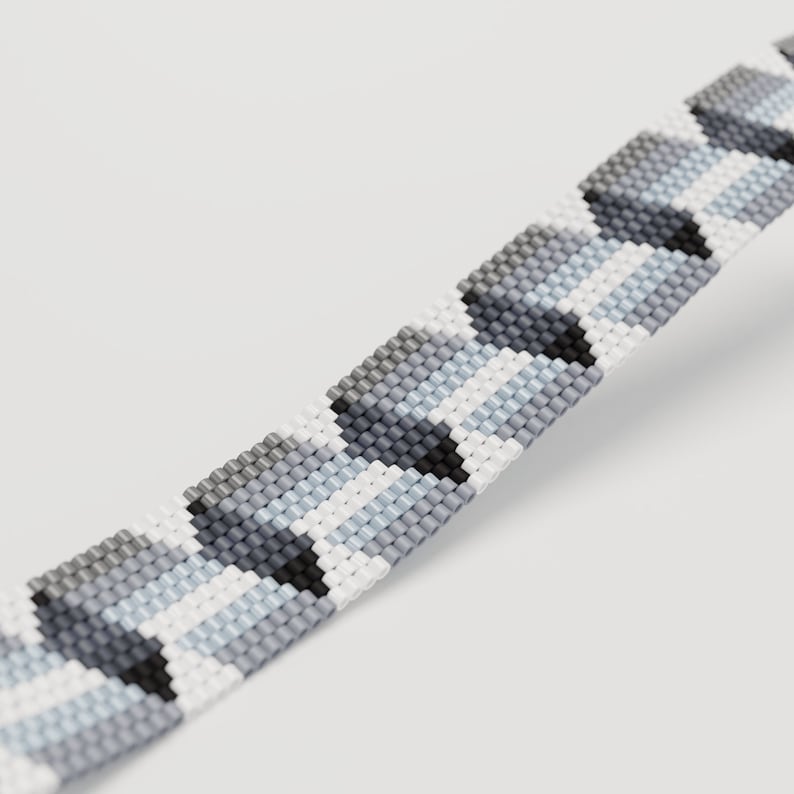 May include: A beaded bracelet featuring a geometric pattern in shades of gray, white, and black. The bracelet is made of small, evenly spaced beads, creating a textured surface. The design is a repeating pattern of shapes.