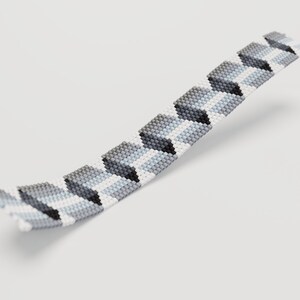 May include: A beaded bracelet with a geometric pattern. The bracelet features a repeating design of gray, black, and white blocks. The beads are small and tightly woven together, creating a textured surface. The bracelet is curved, suggesting it is flexible.