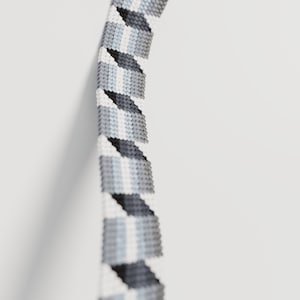 May include: A close-up of a beaded bracelet with a geometric pattern. The bracelet features a repeating design of gray, white, and light blue triangles. The beads are tightly woven together, creating a textured surface against a white background.
