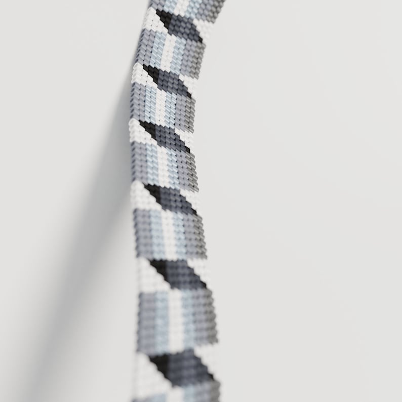 May include: A close-up of a beaded bracelet with a geometric pattern. The bracelet features a repeating design of gray, white, and light blue triangles. The beads are tightly woven together, creating a textured surface against a white background.