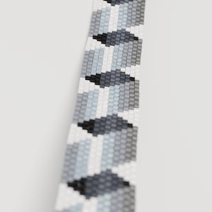 May include: Close-up of a beaded bracelet with a geometric pattern. The bracelet features a repeating design of black, white, and various shades of gray beads. The beads are small and square, creating a textured surface. The background is plain white.