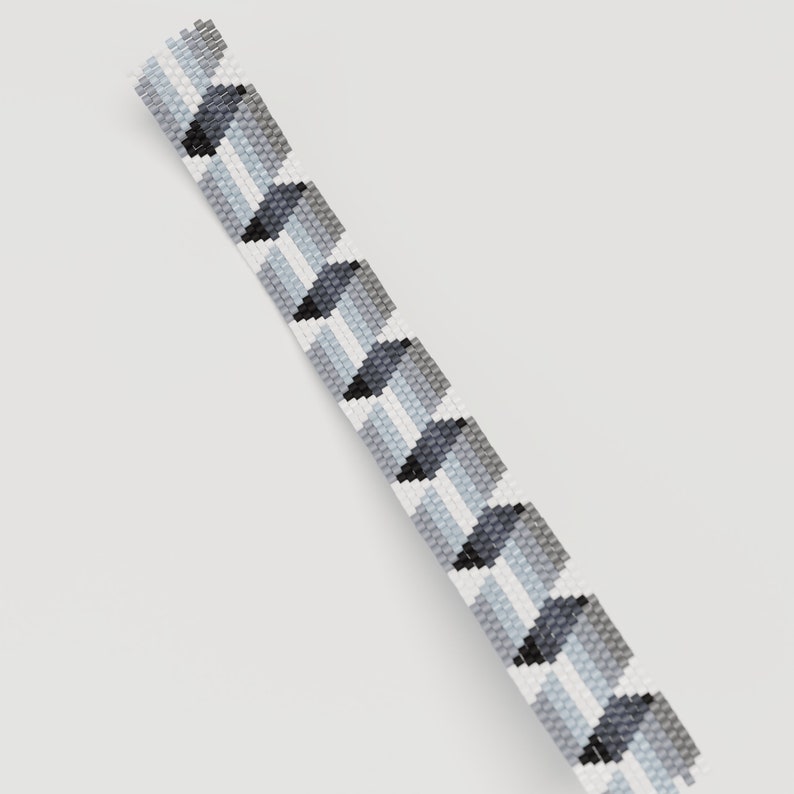 May include: A beaded bracelet with a geometric pattern. The bracelet features a repeating design of gray, black, and white squares. The beads are small and evenly spaced, creating a textured surface. The bracelet is shown against a white background.
