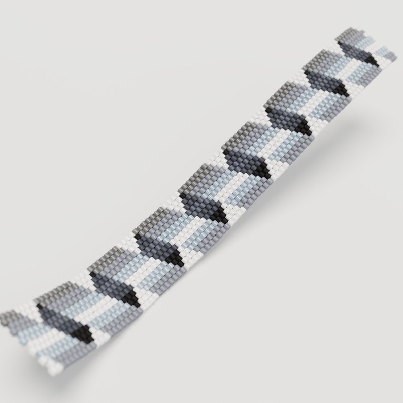 May include: A beaded bracelet featuring a geometric pattern in shades of gray, white, and black. The bracelet is made of small, evenly spaced beads, creating a textured surface. The design has a repeating chevron-like pattern.