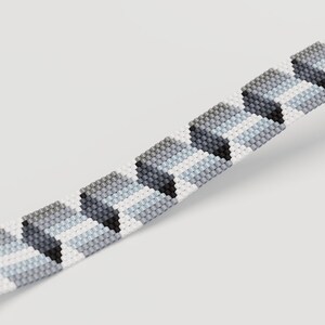 May include: Close-up of a beaded bracelet with a geometric pattern. The bracelet features a repeating design of gray, black, and white beads. The beads are arranged in a chevron pattern, creating a textured look. The bracelet is displayed against a white background.