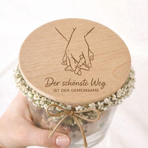 Könnte beinhalten: Ein klares Glas mit einem Holzdeckel mit einem eingravierten Design von zwei Händen, die sich halten. Der Deckel trägt den Text "Der schönste Weg ist der gemeinsame". Das Glas ist mit kleinen weißen Blumen verziert und mit einer Schnur gebunden.