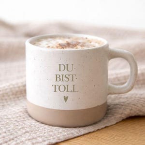 Könnte beinhalten: Eine weiße Keramiktasse mit beigem Boden, gefüllt mit Cappuccino. Auf der Tasse steht in hellgrauer Schrift "DU BIST TOLL", darunter ein kleines Herzsymbol. Die Tasse steht auf einer beigen Oberfläche.