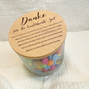 May include: A clear glass jar filled with colorful candies, topped with a wooden lid. The lid has engraved text in German. The jar sits on a white, textured surface.