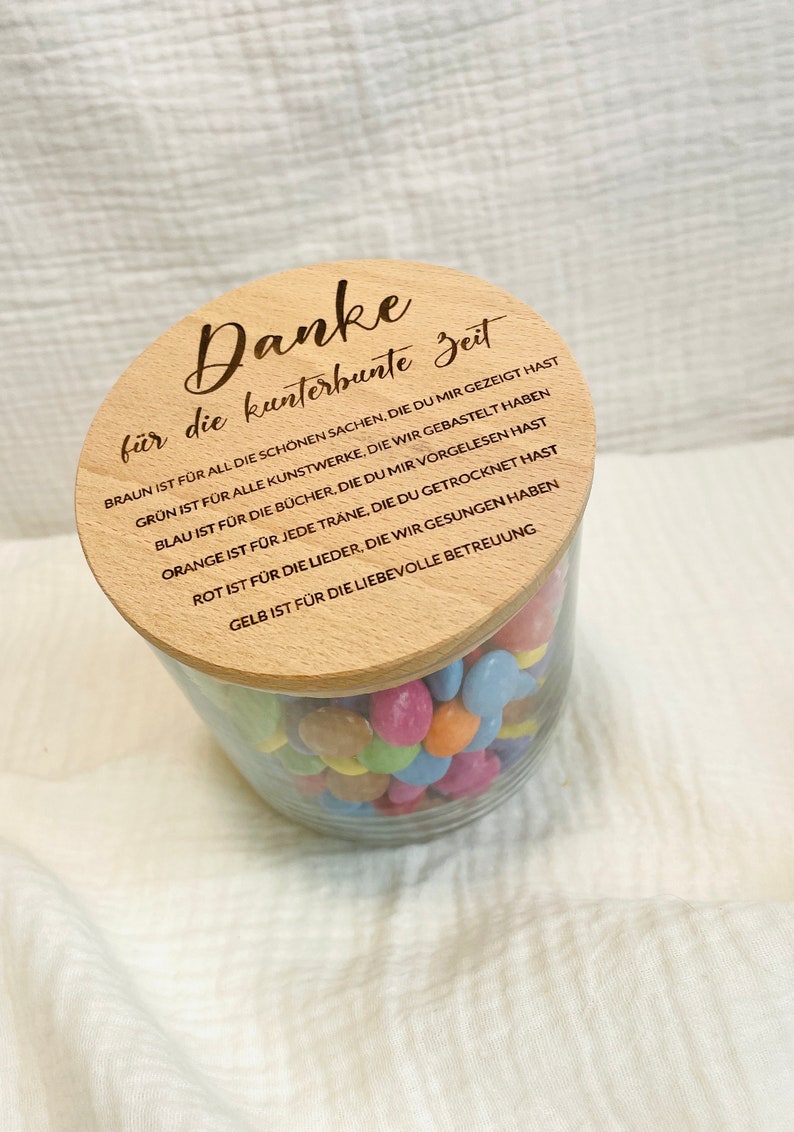 May include: A clear glass jar filled with colorful candies, topped with a wooden lid. The lid has engraved text in German. The jar sits on a white, textured surface.
