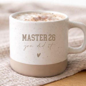 Könnte beinhalten: Eine weiße Keramiktasse mit beigem Boden, gefüllt mit Cappuccino. Auf der Tasse steht der Text "MASTER 26 you did it" und ein Herzsymbol. Die Tasse hat einen Henkel.