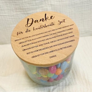 May include: A clear glass jar filled with colorful candies, topped with a wooden lid. The lid has engraved text in German. The jar is filled with various colors of candies.