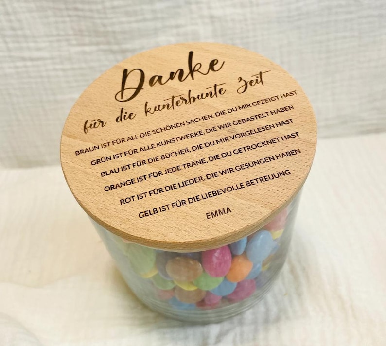 May include: A clear glass jar filled with colorful candies, topped with a wooden lid. The lid has engraved text in German. The jar is filled with various colors of candies.