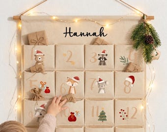May include: A beige fabric wall-hanging advent calendar with twelve pockets, each numbered and decorated with illustrations of woodland animals in Santa hats, gifts, and greenery. The name Hannah is printed in black script at the top.