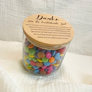 May include: A clear glass jar filled with colorful candies. The jar has a wooden lid with the German text "Danke f&uuml;r die kunterbunte Zeit" and other text. The candies are various colors, including brown, pink, orange, and blue.