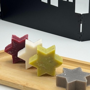 Può includere: Un vassoio di legno contiene quattro saponi a forma di stella nei colori bordeaux, bianco, verde e grigio. I saponi sono disposti in fila, con una forma geometrica nera sullo sfondo.