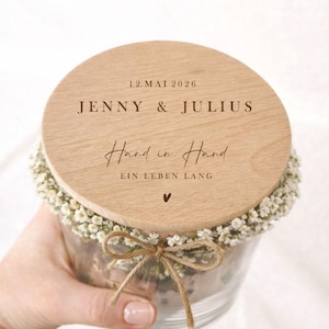 May include: A clear glass jar with a wooden lid, decorated with small white flowers and a twine bow. The lid is engraved with the names "JENNY & JULIUS", the date "12. MAI 2026", and the phrase "Hand in Hand EIN LEBEN LANG".