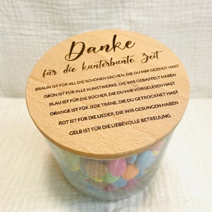 May include: A clear glass jar filled with colorful candies, topped with a wooden lid. The lid is engraved with the German phrase "Danke f&uuml;r die kunterbunte Zeit" and other text. The jar is sitting on a white surface.