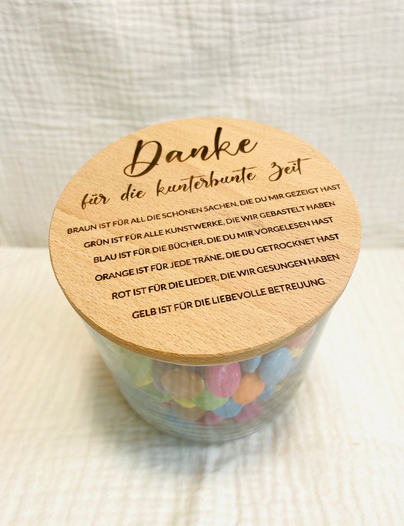 May include: A clear glass jar filled with colorful candies, topped with a wooden lid. The lid is engraved with the German phrase "Danke f&uuml;r die kunterbunte Zeit" and other text. The jar is sitting on a white surface.