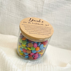 May include: A clear glass jar filled with colorful, round candies. The jar has a wooden lid with German text. The candies are various colors, including blue, green, pink, and yellow. The jar sits on a white, textured surface.