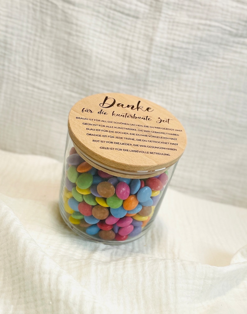 May include: A clear glass jar filled with colorful, round candies. The jar has a wooden lid with German text. The candies are various colors, including blue, green, pink, and yellow. The jar sits on a white, textured surface.