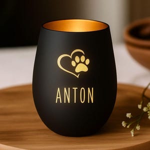Könnte beinhalten: Ein schwarzes Weinglas ohne Stiel mit goldener Innenseite. Das Glas hat ein goldenes Herz- und Pfotenabdruck-Design mit dem Namen "ANTON" darunter. Das Glas steht auf einem Holztablett.