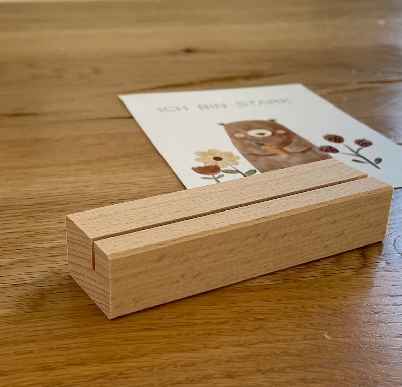 K&ouml;nnte beinhalten: Ein Holz-Kartenhalter mit zwei Schlitzen zum Anzeigen von Karten. Der Halter ist aus hellem braunem Holz gefertigt und hat eine nat&uuml;rliche Oberfl&auml;che. Der Kartenhalter steht auf einer Holzoberfl&auml;che.
