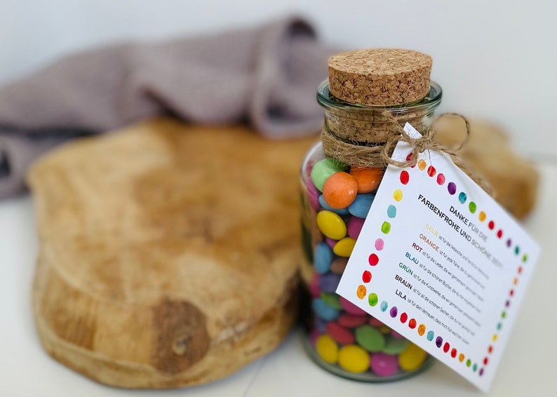 May include: A glass jar filled with colorful candy, tied with twine and a cork lid. A card attached to the jar reads "Danke f&uuml;r die Farbenfrohe und sch&ouml;ne Zeit" with a list of colors and their corresponding number of candies.
