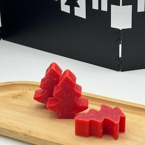Può includere: Tre candele profumate rosse a forma di albero di Natale su un vassoio di legno. Le candele sono di un rosso vibrante e hanno una superficie strutturata. Uno sfondo nero geometrico è visibile dietro il vassoio.