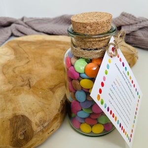 May include: A glass jar filled with colorful candy coated chocolate pieces. The jar has a cork lid and is tied with twine. A white tag is attached to the jar with the text "Danke f&uuml;r die Farbenfrohe und sch&ouml;ne Zeit" and a list of colors in German.