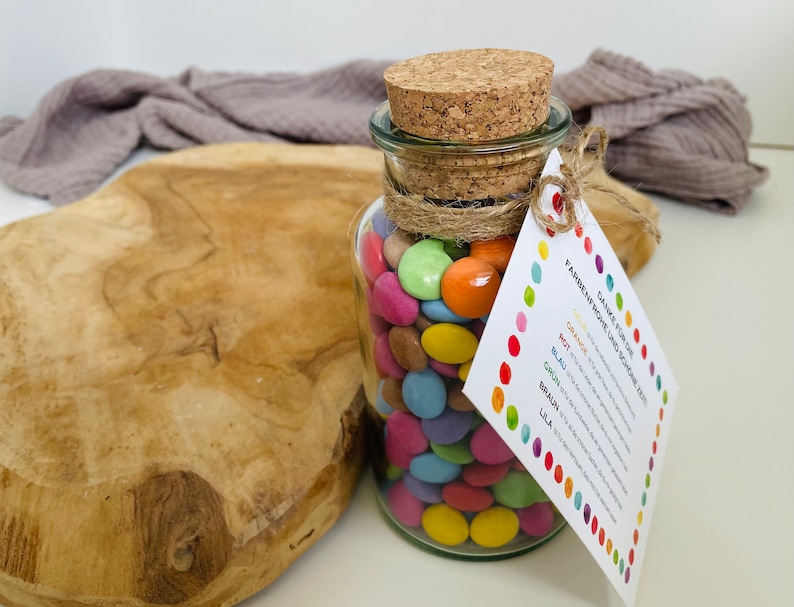 May include: A glass jar filled with colorful candy coated chocolate pieces. The jar has a cork lid and is tied with twine. A white tag is attached to the jar with the text "Danke f&uuml;r die Farbenfrohe und sch&ouml;ne Zeit" and a list of colors in German.
