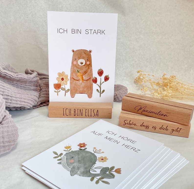K&ouml;nnte beinhalten: Ein Satz Holz-Karten mit Illustrationen eines B&auml;ren, eines Wals und Blumen. Die Karten haben deutschen Text darauf, darunter "Ich bin stark", "Ich bin Elisa" und "Ich h&ouml;re auf mein Herz". Die Karten sind auf einem Holzst&auml;nder gestapelt, auf dem der Name "Maximilian" eingraviert ist.