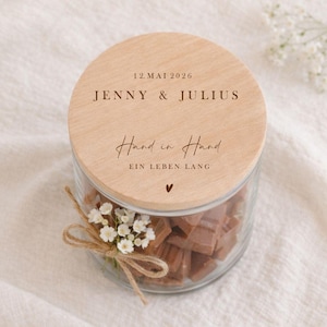 May include: A glass jar filled with brown sweets, with a wooden lid. The lid is engraved with the names "JENNY & JULIUS" and the date "12. MAI 2026". The words "Hand in Hand" and "EIN LEBEN LANG" are also engraved on the lid.