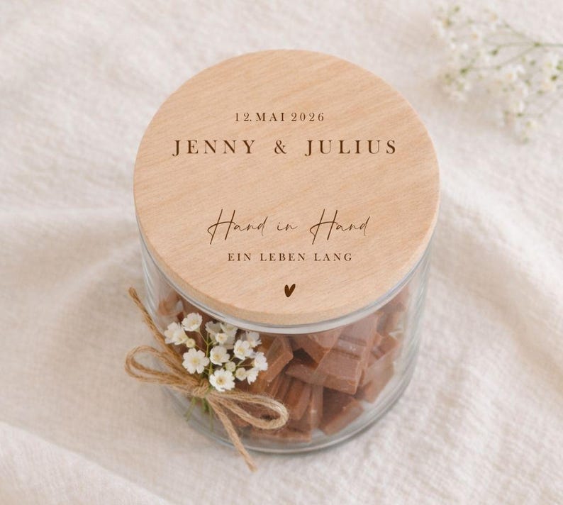 May include: A glass jar filled with brown sweets, with a wooden lid. The lid is engraved with the names "JENNY & JULIUS" and the date "12. MAI 2026". The words "Hand in Hand" and "EIN LEBEN LANG" are also engraved on the lid.