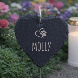 Könnte beinhalten: Ein herzförmiger, dunkelgrauer Gedenkpfahl für Haustiere aus Schiefer mit dem Namen "MOLLY" in weißen Buchstaben. Ein Pfotenabdruck-Design befindet sich über dem Namen. Der Pfahl befindet sich in einem Garten mit Blumen und einer brennenden Kerze im Hintergrund.