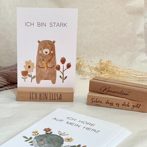 K&ouml;nnte beinhalten: Ein Satz illustrierter Karten mit positiven Affirmationen auf Deutsch. Die Karten zeigen einen B&auml;ren, der eine Karotte h&auml;lt, einen Wal und Blumen. Die Karten werden auf Holzst&auml;ndern mit dem Text "Ich bin stark", "Ich bin Elisa", "Maximilian", "Sch&ouml;n, dass es dich gibt" und "Ich h&ouml;re auf mein Herz" angezeigt.