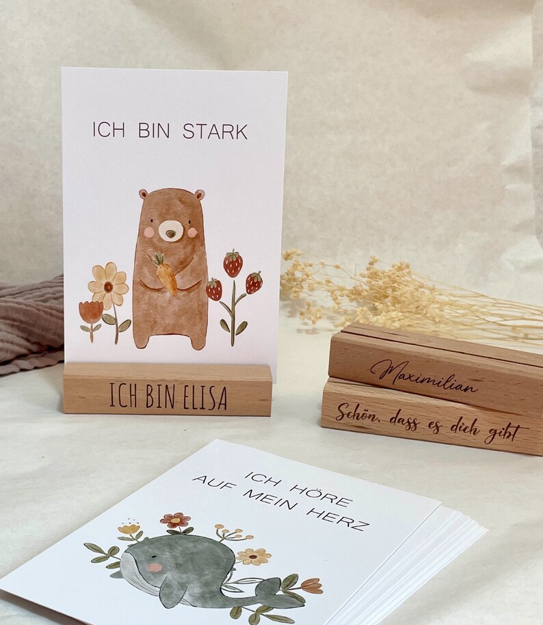 K&ouml;nnte beinhalten: Ein Satz illustrierter Karten mit positiven Affirmationen auf Deutsch. Die Karten zeigen einen B&auml;ren, der eine Karotte h&auml;lt, einen Wal und Blumen. Die Karten werden auf Holzst&auml;ndern mit dem Text "Ich bin stark", "Ich bin Elisa", "Maximilian", "Sch&ouml;n, dass es dich gibt" und "Ich h&ouml;re auf mein Herz" angezeigt.