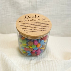 May include: A clear glass jar filled with colorful candies, topped with a wooden lid. The lid has engraved text in German. The jar is sitting on a white surface.