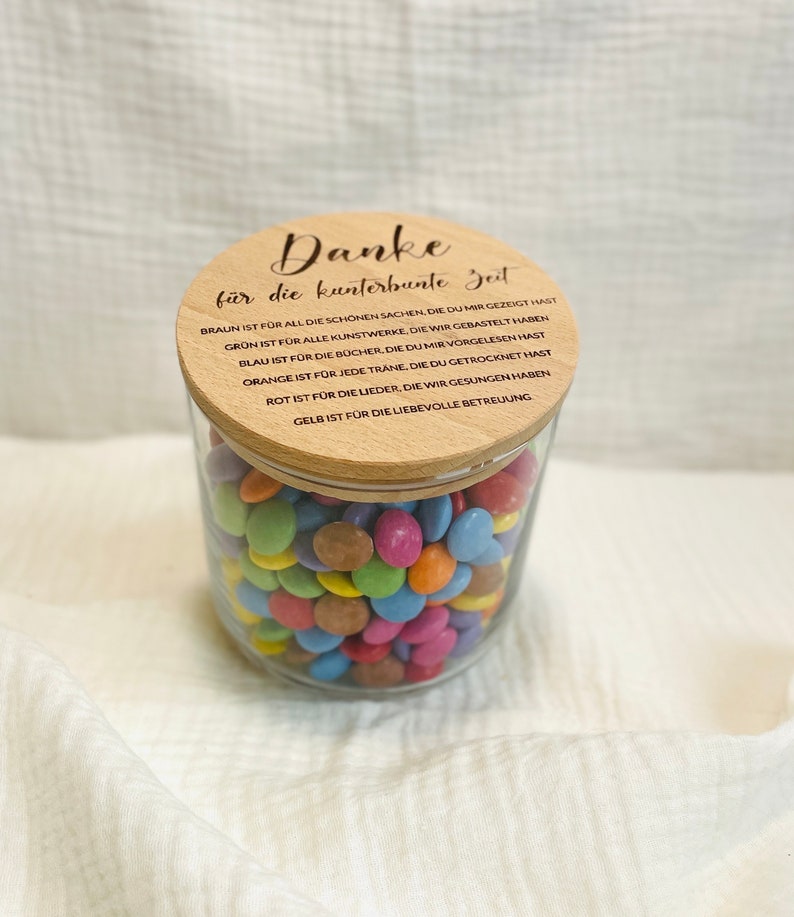 May include: A clear glass jar filled with colorful candies, topped with a wooden lid. The lid has engraved text in German. The jar is sitting on a white surface.