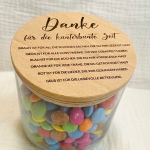 May include: A clear glass jar filled with colorful candies, topped with a wooden lid. The lid has engraved text in German. The jar contains a variety of candies in shades of blue, green, orange, pink, and yellow.