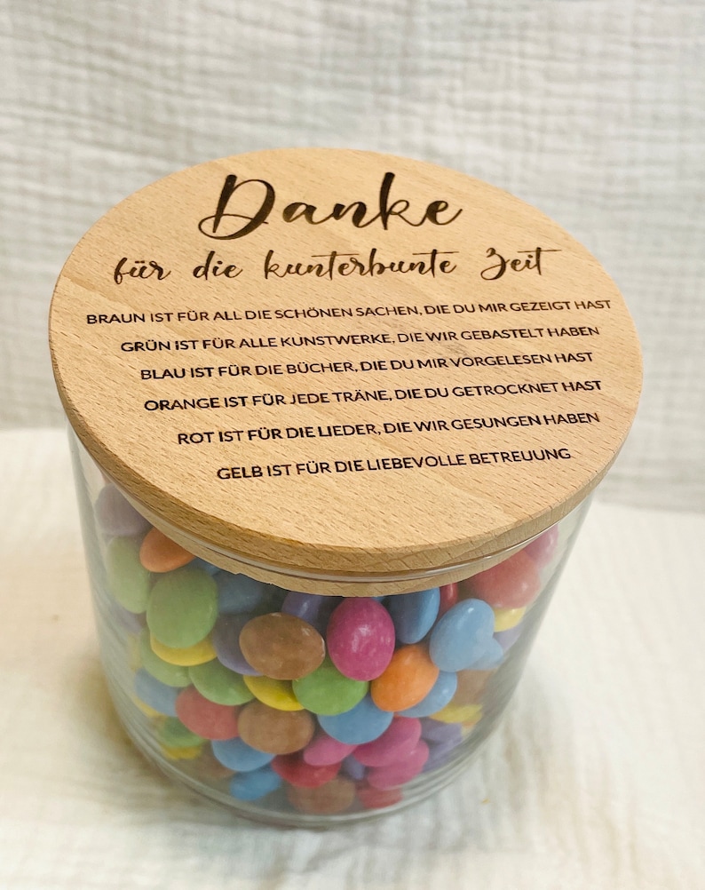 May include: A clear glass jar filled with colorful candies, topped with a wooden lid. The lid has engraved text in German. The jar contains a variety of candies in shades of blue, green, orange, pink, and yellow.