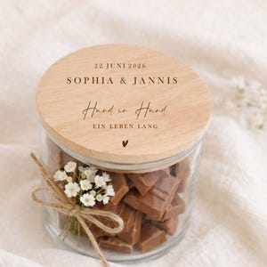 May include: A glass jar filled with chocolate pieces, topped with a wooden lid. The lid is engraved with the names "SOPHIA & JANNIS", the date "22. JUNI 2026", and the phrase "Hand in Hand EIN LEBEN LANG". A small bouquet of white flowers is tied around the jar.