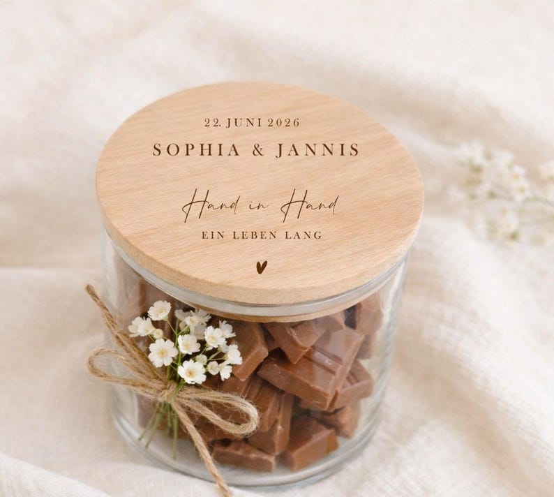 May include: A glass jar filled with chocolate pieces, topped with a wooden lid. The lid is engraved with the names "SOPHIA & JANNIS", the date "22. JUNI 2026", and the phrase "Hand in Hand EIN LEBEN LANG". A small bouquet of white flowers is tied around the jar.
