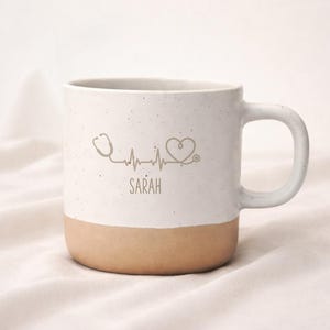 Könnte beinhalten: Ein weißer Keramikbecher mit gesprenkelter Oberfläche und beigem Boden. Auf der Vorderseite befindet sich eine braune Grafik eines Stethoskops, das eine Herzschlaglinie bildet, die in ein Herz übergeht, mit dem Namen SARAH in braunen Großbuchstaben.
