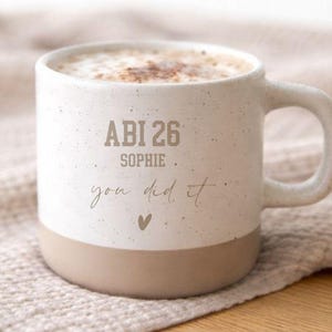 Könnte beinhalten: Eine weiße Keramik-Kaffeetasse mit gesprenkelter Oberfläche, mit einem beigefarbenen Boden und Henkel. Die Tasse ist mit einem Cappuccino gefüllt und trägt den Text "ABI 26 SOPHIE you did it" auf der Vorderseite.